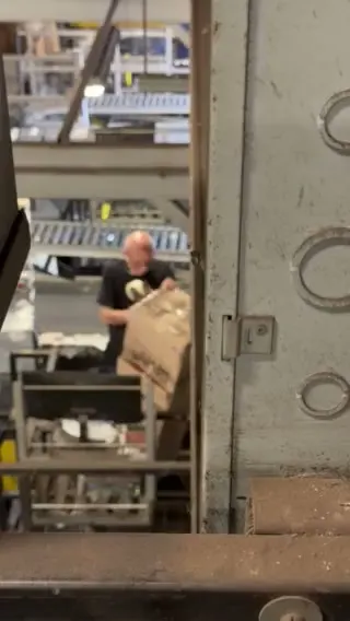 Seen through a gap between steel columns, a hub worker lifts a torn carton off a cart beside a conveyor.